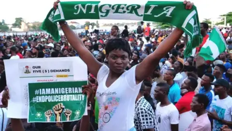 Getty Images A woman in a white T-shirt stands in front of a crowd of people. She is holding a green-and-white scarf above her head, which says "Nigeria".
