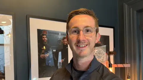 Peter McAuley has short brown hair and glasses and is staring into the camera. He is wearing a dark jacket with a dark top underneath. In the background is a picture on a wall and dark curtains.