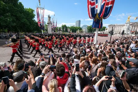 Getty Images Crowds line both sides of the Mall and watch musicians from the Massed Bands of the Household Division parade from Buckingham Palace. Rows of onlookers stand in the sunshine, many with their mobile phones aloft. On the Mall, musicians in red, white and black uniform parade and play their instruments.