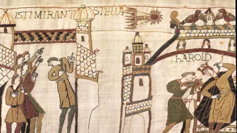 Bayeux Museum A wide crop shows the scene of people looking up at the sky and pointing while a comet flies above, they are surrounded by medieval buildings, and at the top of the tapestry you can read numbers 31, 32 and 33.