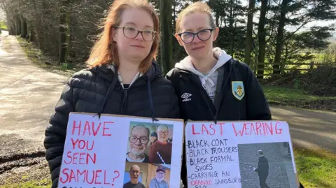 Olivia Richwald/BBC Two sisters hold handwritten signs attached around their necks. One says "have you seen Samuel"? with four photos of an older man. The other has a bullet point list describing the clothes he was last seen in, next to a CCTV image of him.