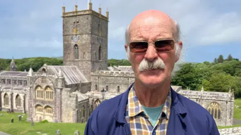Malcolm standing in front of St Davids Cathedral on a sunny day. He wears a blue t-shirt, yellow chequerd shirt and a blue jacket. Has brown sunglasses and has short grey hair. 