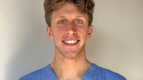 Sam Gurling Sam wearing light blue medical scrubs stands indoors against a plain, light‑coloured wall