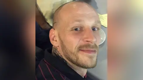 Close up of Callum Kerr with a shaved head and light brown beard wearing a black and pink striped polo top. He is smiling. There is a table and white plate in the background. 