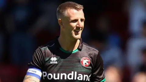 Peter Clarke wearing a black Walsall football top while on the pitch. He is squinting and smiling. He is looking away from the camera.