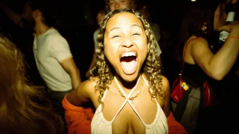 3flavourscornetto Uma mulher abre bem a boca enquanto olha diretamente para a câmera enquanto dança. Ela tem cabelos castanhos na altura dos ombros e usa uma blusa branca.