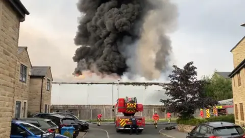 Thick black smoke rises in the air from the large grey industrial unit. A fire engine is parked at the end of Knoll Close which is a residential street of two-storey, almost identical beige brick buildings. There are lots of parked cars and three firefighters in hi-vis orange jackets, yellow trousers and white helmets are walking around the engine. A fourth firefighter in black overalls stands behind the engine which has a large ladder on it.