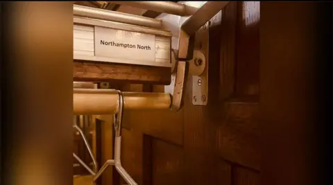 Lucy Rigby Wooden rail with coat hanger attached and with "Northampton North" label above it