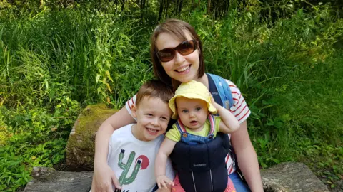 Darren Wheatley Nicola with Oscar and Ffion. Ffion is a baby in a yellow sun hat and t-shift being held in a baby carrier. Oscar has brown hair and is wearing a white t-shirt with a cactus on it. Nicola has a brown bob haircut and is wearing sunglasses. All are smiling.