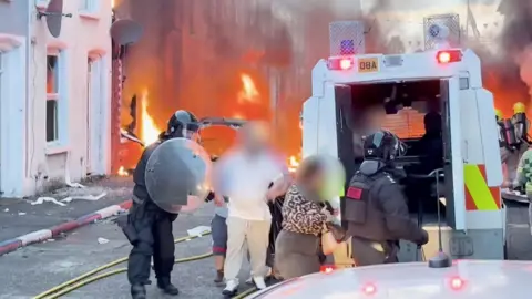 People with their faces blurred being rushed into a van by police as fire burns near their home.