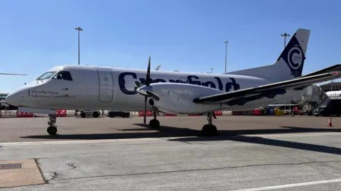 Nicola Haseler/BBC A side view of the National Flying Laboratory Centre - a grey aircraft packed with scientific equipment. The photo was taken on a runway on a bright sunny day. 