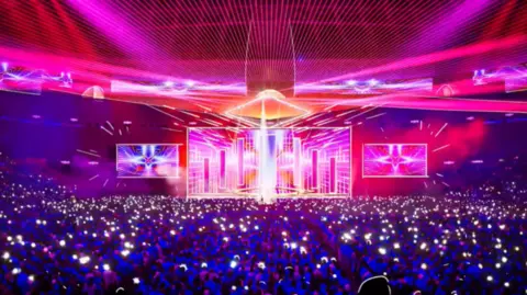 BCFC Forum A concert setting, with screens at the front and purple and red lighting. Many people watching are holding a light. 