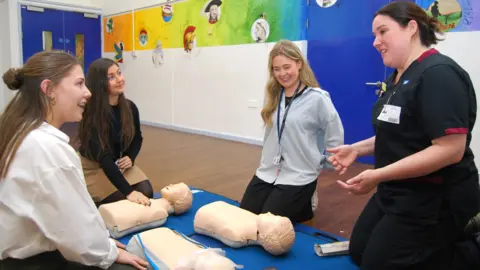 KCH Four people in a room, they are all kneeling. A woman, dressed as a nurse in a dark top and trousers, with her hands out, talking to three other women, with three resuscitation dummies in front of them, on a blue map. There is artwork on the walls and a blue door to the left. 
