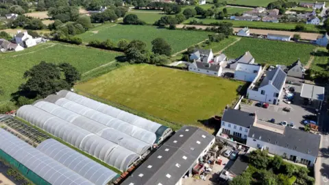 An aerial view of a rural area featuring several greenhouses with curved roofs, a large grassy field, and multiple buildings including residential houses and farm structures. The landscape is lush with greenery, trees, and cultivated fields. The buildings are arranged in clusters with some vehicles parked near them. The scene is set in a countryside environment with clear skies.