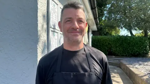 A man standing in front of a white building. He is smiling and wearing a black t-shirt and apron. There is a green hedge behind him and tall trees. 