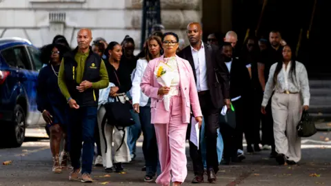 PA Media A group of people outside a court building, a woman at the front in a pink suit, holding a bunch of flowers.