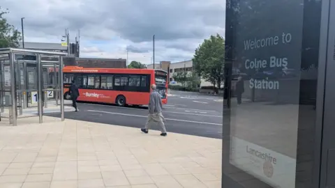 A view of the new bus station with a large flagged area in the foreground and large asphalt area with a red single decker bus driving past glass and chrome bus shelters. To the right is a digital sign saying 'welcome to Colne bus station'