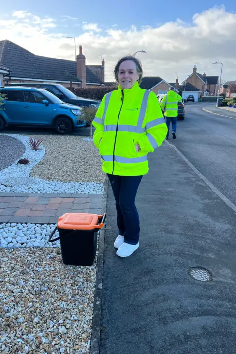 A woman with bobbed hair wearing a hi-vis jacket stands next to a black and orange food waste caddie ready for collection outside a house