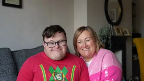 Joel and Clare smiles at the camera, Joel is wearing a red jumper with a green samurai character on the front. He has short brown hair and is wearing black framed glasses. Clare is sat close to him, she is wearing a two-tone pink jumper and short shoulder-length blond hair. They are sat in their living room on a sofa. 