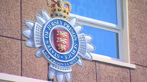A close up of a sign for Jersey's only prison. The crest is blue with a red shield in the middle with three lions on it. There is also a crown sitting on the blue crest.