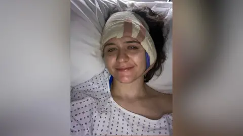 Suzy Spens Suzi in a hospital bed. She smiled on camera and is wearing a hospital gown with a bandage around her head.