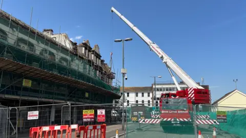 A large crane is lifting workmen to work on the building. The crane is behind security fencing with warning signs of a demolition site.