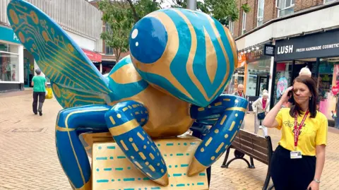 Steve Huntley/BBC A giant bee sculpture painted in blue and gold stands in a pedestrianised street, close to a bench and shops. A female charity worker in a yellow Buzz in the City T-shirt is standing next to it.