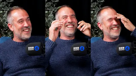 Dr Xand van Tulleken in a navy blue jumper on the What's Up Docs? podcast. He is laughing while speaking into a black mic that has the words 'BBC Radio 4' on it.