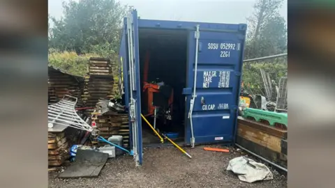 The Treeple The picture shows the bent metal on the front of the broken into container unit. The container is blue and some scattered tools are visible inside. 