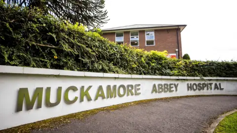 PA Media Una pared blanca dice 'Muckamore Abbey'. Hay un edificio de ladrillo rojo detrás de la pared baja. Un pequeño arbusto verde corre a lo largo de la parte superior de la pared blanca. 