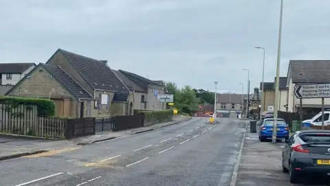 A general view of Bathgate Road in Blackburn, West Lothian. There are cars parked on the right of the street. On both sides, there is pavement. A crossing island is in the background of the image.