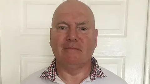 Philip Miller, who has a bald head and is wearing a white shirt with a dark red and black checked collar, is standing in front of a white door in a house. He looks impassive. 