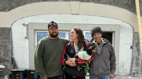 Ellie Pizey Joe, Ellie and Simon are standing together outside the building. She is smiling at Joe while Joe and Simon are smiling at the camera. Ellie is holding a small black and tan dog in her arms which is wearing a coat. Joe has a baseball cap on and Simon a woolly hat. Behind them is a stack of plastic boxes which hold tools and equipment. There is a piece of wood leaning against the building which has a large bifold glass door.