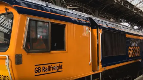The engine at the station. It has a bright yellow cab with a navy blue roof and says "GB Railfreight" under the driver's side window. The first carriage is navy blue with the number 66303 in yellow under the plaque