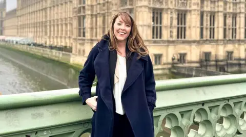 A woman with brown hair wearing a white shirt under a navy trench coat. She is standing in front of the Houses of Parliament.