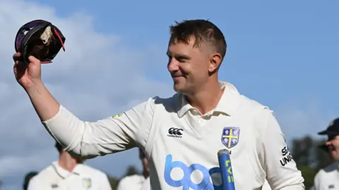 Scott Borthwick holding his cap aloft over his head while smiling off-camera.