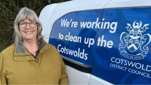 Councillor Juliet Layton in a yellow/brown coat looking into the camera. She is standing next to a branded Cotswold District Council van.