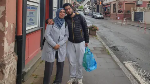 Family Missing Ali Durrani with his mother Mahjabeen. The pair are on an old-town looking street and posing for a photo. She is wearing a headscarf and a long cardigan. He has on a black outdoor jacket.
