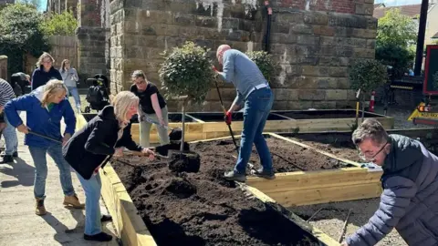 Nine men and women using gardening tools to fill raised wooden flower beds with soil. They are also adding plants. There is an old stone building in the background.
