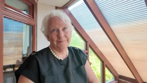 A woman with white hair in a black dress is standing on the balcony of the Oxford eco home with a sloped glass roof behind her