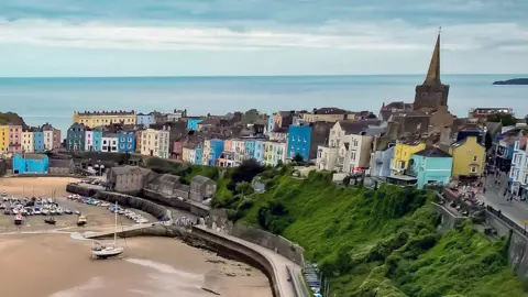 Aerial photo of the seaside town of Tenby in Pembrokeshire, Wales, showing rows of colourful houses overlooking the harbour with boats on the sand as the tide is out, with the Atlantic Ocean in the background behind the town's church spire and some clouds in the sky.