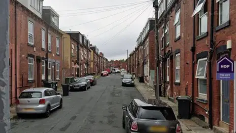 A residential street with back-to-back redbrick properties on both sides of the street. Parked cars and bins can also be seen.
