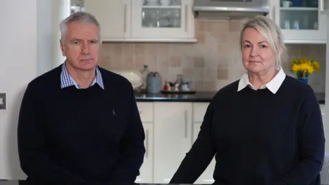 A couple wearing shirts and dark sweatshirts, sat in a kitchen