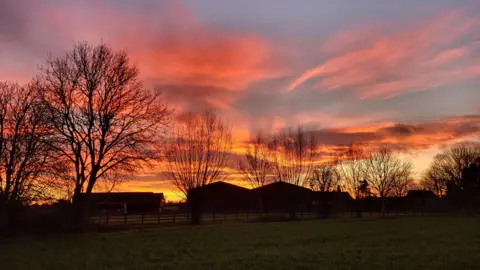 The sky is a mixture of yellow, orange and pink. Beneath it sits a green field and wooden fence with several large farm buildings behind the fence.