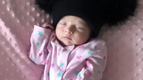 A baby wearing a pink babygrow with blue stars and dark pint hears on it. She is asleep while lying on a pink blanket and wearing a black hat