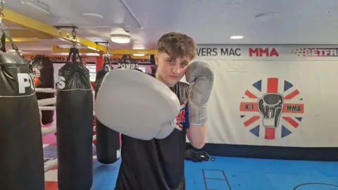 BBC/Naj Modak Luke wearing boxing gloves training in a gym, standing in front of hanging punch bags and a ‘Powers Mac MMA’ banner.”