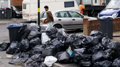 A large pile of black bin bags in the street with a man walking behind them and a white caravan parked on the opposite side of the road