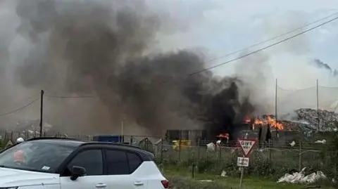 A view of a large smoke plume in the sky, flames at the bottom on large piles of rubbish. There are fences, a road and a couple of cars in the foreground.