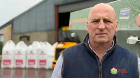 A man standing in a farm. He is wearing a blue shirt and has a navy body warmer with the words Ulster Farmers Union written on it