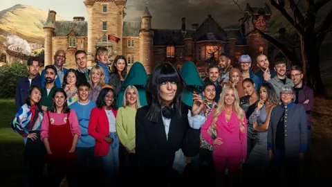 BBC/Studio Lambert Claudia Winkleman berdiri di depan 25 kontestan dengan latar belakang kastil/rumah megah Skotlandia dalam foto publisitas untuk The Traitors. Claudia melontarkan pandangan aneh dari balik poni khasnya. Dia mengenakan jaket hitam di atas blus putih, dengan manset lebar yang menonjol dari ujung lengan.